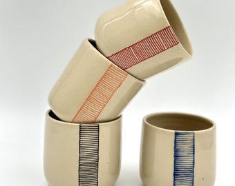 May include: Four ceramic mugs with different colored stripes. The mugs are stacked on top of each other, with the top mug tilted to the side. The stripes are red, orange, blue, and black.