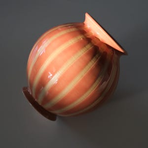 May include: A peach-colored ceramic vase with a rounded body and a flared, angled rim. The vase features vertical stripes in a lighter, cream color. The base is a darker shade of peach. The vase is likely for decorative purposes.