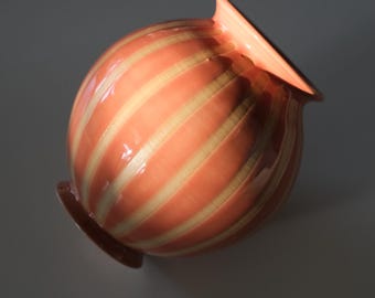 May include: A peach-colored ceramic vase with a rounded body and a flared, angled rim. The vase features vertical stripes in a lighter, cream color. The base is a darker shade of peach. The vase is likely for decorative purposes.