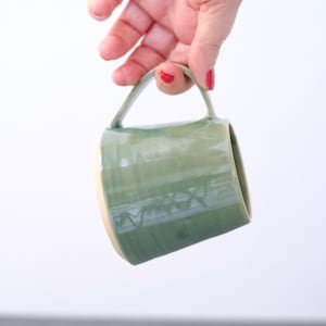 Könnte beinhalten: Ein Keramikbecher mit einer glänzenden salbeigrünen Glasur und einem glatten, konisch zulaufenden zylindrischen Körper. Der Becher verfügt über einen einzelnen eckigen Henkel an der Seite und einen hellen Bodenrand, der sich von der grünen Oberfläche abhebt.