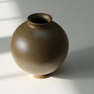 May include: A round, brown ceramic vase with a narrow neck and a wide, spherical body. The vase has a speckled texture and sits on a small, circular base. The interior of the vase is dark, and the lighting creates shadows.