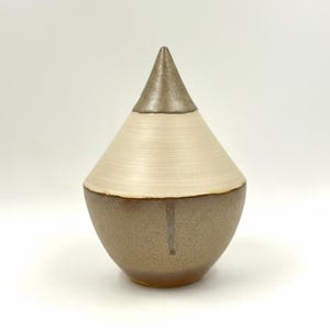 May include: A handcrafted ceramic vase with a unique, teardrop shape. The vase features a metallic bronze top, a cream-coloured middle section, and a bronze base. The design is simple yet elegant, suitable for home decor.