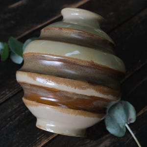 May include: A handmade ceramic vase with a unique, ridged design. The vase features alternating bands of light beige and brown, creating a spiral effect. The vase has a rounded base and a narrow opening at the top, perfect for displaying flowers.