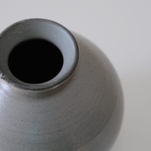 May include: A small, round, ceramic vase in a muted gray color. The vase has a wide opening and a slightly textured surface. The interior is dark, contrasting with the light exterior. A simple, minimalist design.