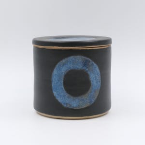 May include: A cylindrical ceramic jar with a matte black finish and a matching lid. The front of the jar features a prominent, textured circular design in shades of blue. The base and rim of the jar reveal a natural, unglazed tan clay edge.