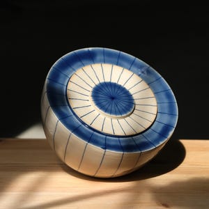 Könnte beinhalten: Eine weiße Keramikschüssel mit blau-weißem Streifenmuster. Die Schüssel hat einen Deckel mit ähnlichem Design. Die Schüssel steht auf einer Holzoberfläche.