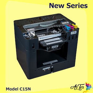 Può includere: Una stampante 3D nera e argentata con un coperchio trasparente e un vassoio per cartucce a colori. La stampante ha un'etichetta che dice "Nuova Serie" e "Modello C15N".