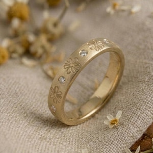 May include: A gold ring featuring a floral design and small diamonds. The ring is displayed on a beige linen cloth, with dried flowers around it. The ring's surface has engraved flowers and small, clear gemstones.