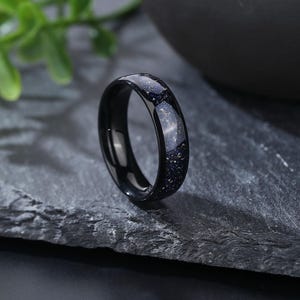 Puede incluir: Un anillo negro con una incrustaci&oacute;n moteada azul y dorada. El anillo est&aacute; hecho de carburo de tungsteno y es una opci&oacute;n popular para las alianzas de boda.