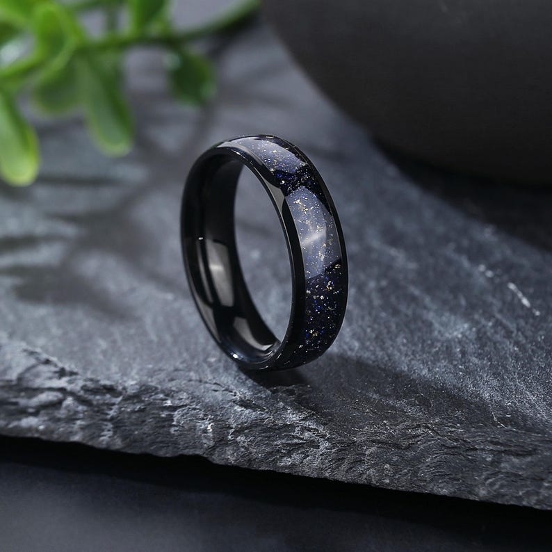 Puede incluir: Un anillo negro con una incrustaci&oacute;n moteada azul y dorada. El anillo est&aacute; hecho de carburo de tungsteno y es una opci&oacute;n popular para las alianzas de boda.