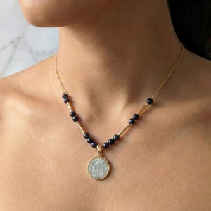 Puede incluir: Collar dorado con un colgante circular con diseño geométrico. La cadena es fina, con cuentas azul oscuro y barras doradas. El colgante es de color azul claro con un borde dorado.
