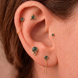 Könnte beinhalten: Ein Set aus goldfarbenen Ohrringen mit runden grünen Moosachat-Steinen. Die Kollektion umfasst einen Ohrstecker mit zarter Kette, einen Stecker mit drei Kügelchen, einen kleinen Einzelstecker, einen Stecker mit größerem Stein und einen mit Kette.