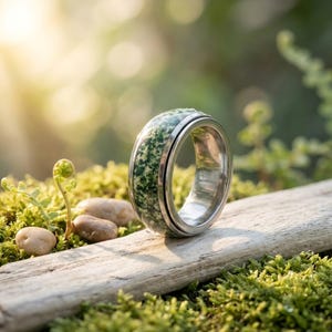 May include: A silver ring with a green and white speckled stone center. The ring is set against a backdrop of green moss, small stones, and a wooden surface, with soft sunlight in the background. The ring's design is modern and natural.