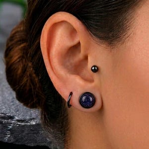 May include: Close-up of an ear with three pieces of jewelry. A small black stud, a black hoop earring, and a large black plug with a dark blue, glittery center. The ear is set against a neutral background.