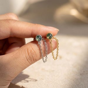 Op de afbeelding: Twee oorknopjes met ronde, in een zetting geplaatste mosagaatstenen met groene insluitingen. De ene oorbel heeft een zilverkleurige metalen afwerking met een fijn kettinkje, de andere een goudkleurige metalen afwerking met een bijpassende ketting.