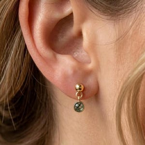 May include: A gold-toned stud earring featuring a small polished sphere at the top and a circular drop pendant below. The pendant contains a round, mottled green and grey stone set within a smooth gold-colored metal bezel frame.