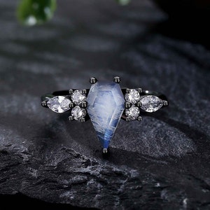 Puede incluir: Un anillo negro con una gran piedra de luna iridiscente engastada en un engaste en forma de ataúd. El anillo está adornado con piedras más pequeñas y transparentes.