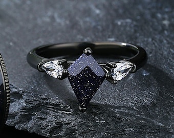 May include: A black ring featuring a diamond-shaped, dark blue stone with sparkling inclusions. Two pear-shaped, clear stones flank the central stone. The ring has a sleek, modern design.