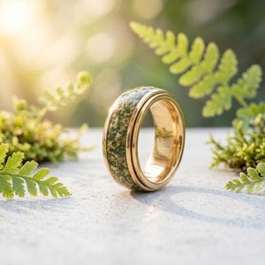Può includere: Un anello color oro con una fascia di pietra verde maculata. L'anello è posto su uno sfondo sfocato di fogliame verde e luce solare, evidenziando il design e la consistenza dell'anello. La fascia dell'anello è larga circa 0,8 cm.