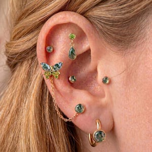 May include: An ear featuring various earrings. A butterfly-shaped earring with green and yellow details is linked by a chain to a round earring. Other earrings include a teardrop, round studs, and a hoop earring, all with green stone features.