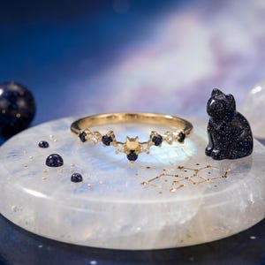 May include: A gold-toned ring with a crescent moon and cat design, adorned with blue and white gemstones. A small, dark blue cat figurine and scattered gemstones are also present, set against a celestial background.