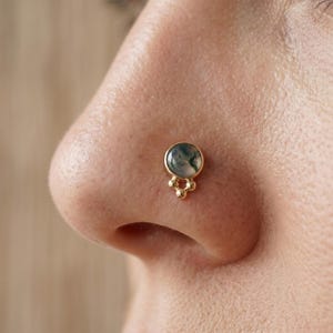 May include: A gold nose stud featuring a round, bezel-set moss agate stone with a translucent, milky appearance and dark green inclusions. The setting is accented with three small, polished gold spheres arranged in a cluster at the base.