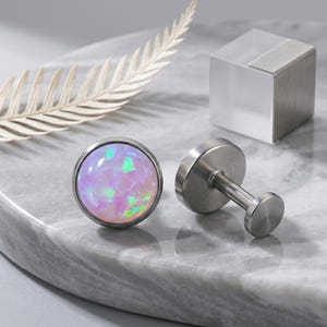 May include: A pair of stainless steel cufflinks with round, iridescent opal stones. The opals display a mix of pink, green, and blue hues. The cufflinks are set on a marble surface.