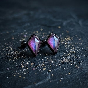 May include: A pair of stud earrings with a diamond-shaped, faceted purple and blue gemstone set in a black metal frame. The earrings are displayed on a dark, textured surface with scattered gold flecks.