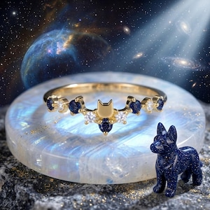 May include: A gold ring with a cat-shaped design, adorned with blue and white gemstones. A small blue dog figurine with glittery specks is placed beside the ring. The backdrop showcases a galaxy.
