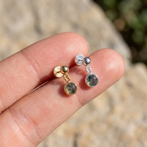 May include: Two stud earrings featuring a small round ball post and a dangling circular charm set with a moss agate stone. One earring is finished in a warm gold tone, while the other is finished in a cool silver tone, both showcasing green inclusions.