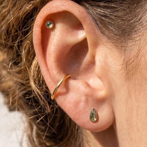 May include: A gold ear cuff, a small round stud earring with a green stone in the upper cartilage, and a teardrop-shaped stud earring with a green stone in the earlobe. The jewelry pieces are made of polished gold and feature natural green stones.