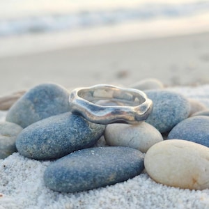Puede incluir: Un anillo plateado con un diseño ondulado y orgánico descansa sobre piedras lisas de color azul grisáceo. La superficie del anillo refleja la luz suave, creando un brillo sutil. El anillo es una joya, probablemente para uso diario.