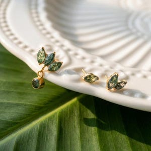 May include: Three gold-tone earrings featuring marquise-cut green moss agate stones arranged in leaf-like clusters. One earring includes a dangling round green stone, while the other two are stud-style earrings with small, pointed green stone accents.