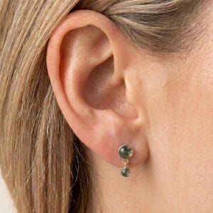 May include: A gold drop earring featuring two round, moss green gemstone cabochons. The earring consists of a larger circular stone at the top connected by a thin gold link to a smaller circular stone dangling below, secured to the earlobe.