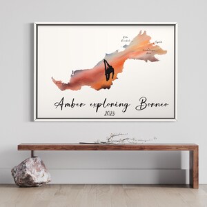 May include: A watercolor painting of the island of Borneo with a silhouette of a person exploring. The painting has a sunset color scheme with orange, red, and yellow hues. The text "Amber exploring Borneo 2023" is written below the map.