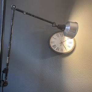 May include: A black adjustable desk lamp with a silver shade is positioned above a round wall clock. The clock has a white face with black Roman numerals and hands. The lamp is mounted on a dark gray wall.