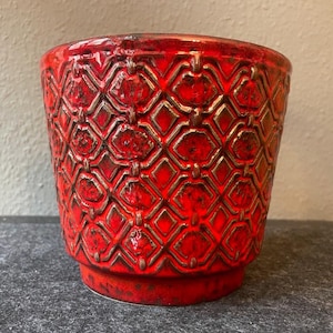 Puede incluir: Una maceta de cerámica roja con un patrón geométrico. La maceta tiene una base ancha y un borde ligeramente acampanado.
