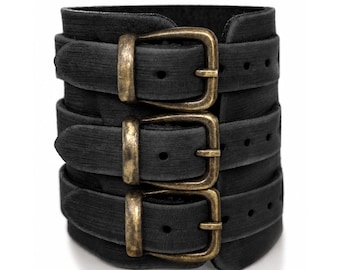 May include: A wide black cuff bracelet featuring three horizontal straps, each secured with a rectangular antique brass-toned metal buckle. The black material has a subtle horizontal textured finish across the surface of the three bands.