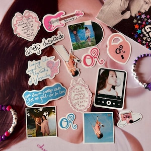Puede incluir: Una colección de pegatinas y accesorios con una guitarra eléctrica rosa, un disco de vinilo y varias imágenes de una persona. Otras pegatinas incluyen texto como "drop dead" y "looking like an angel on the walls of Versailles."