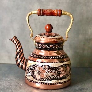 May include: A copper teapot with a wooden handle and intricate floral designs. The teapot has a black and copper striped spout and a gold-colored rim.