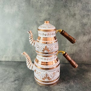 Può includere: Un set da tè turco bianco e rame con intricati disegni floreali. Il set comprende due teiere con manici in legno e un beccuccio.