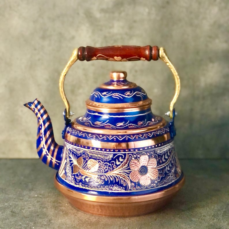 Pode incluir: Uma chaleira azul e cobre com uma al&ccedil;a dourada e desenhos florais intrincados. A chaleira tem um corpo redondo e um bico lateral.