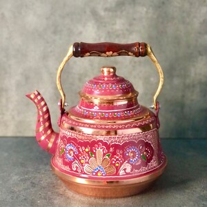 May include: A pink enamel teapot with a copper base and a wooden handle. The teapot is decorated with a floral pattern in shades of pink, blue, green, and gold.