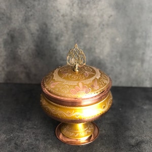 Może przedstawiać: Dekoracyjna miska ze złota i miedzi z ozdobną pokrywką i kwiatowym wzorem. Miska stoi na podstawie i ma mały zwieńczenie na górze pokrywki.