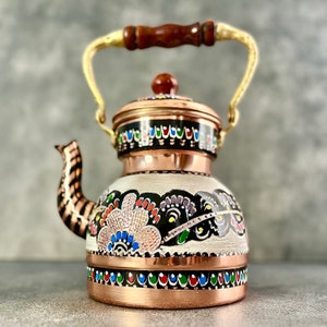 Peut inclure: Une théière en cuivre turque traditionnelle avec un motif floral peint à la main en noir, blanc, rouge, bleu et vert. La théière a une poignée en bois et un couvercle avec un petit bouton.