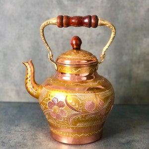 May include: A copper teapot with a decorative floral design etched into the surface. The teapot has a wooden handle and a lid with a small wooden knob. The spout and handle are gold-colored.