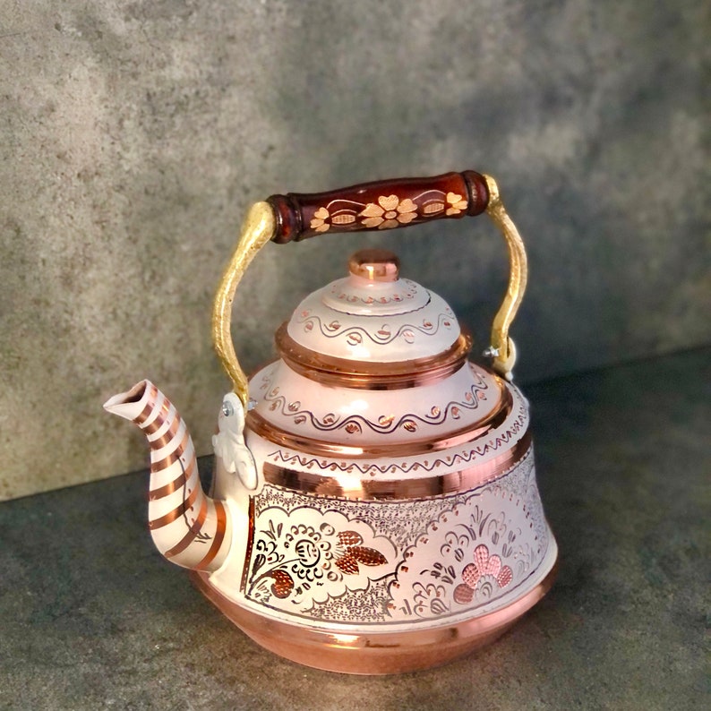 Pode incluir: Uma chaleira branca e de cobre com um design floral. A chaleira tem uma pega de madeira e uma tampa com um bot&atilde;o.