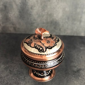 Könnte beinhalten: Eine dekorative Kupferschale mit einem schwarzen und weißen Blumenmuster. Die Schale hat einen Deckel mit einem blattförmigen Griff. Die Schale steht auf einem Sockel.