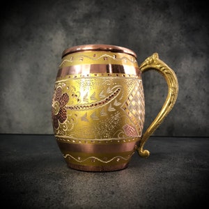 May include: A copper mug with a gold-colored design and a brass handle. The mug has an intricate floral pattern and is perfect for serving beverages.