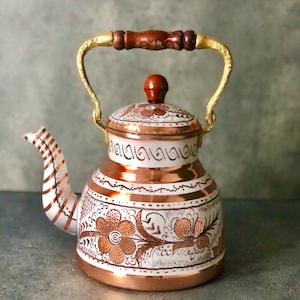 May include: A copper teapot with a white floral design and a wooden handle. The teapot has a gold-colored spout and lid.
